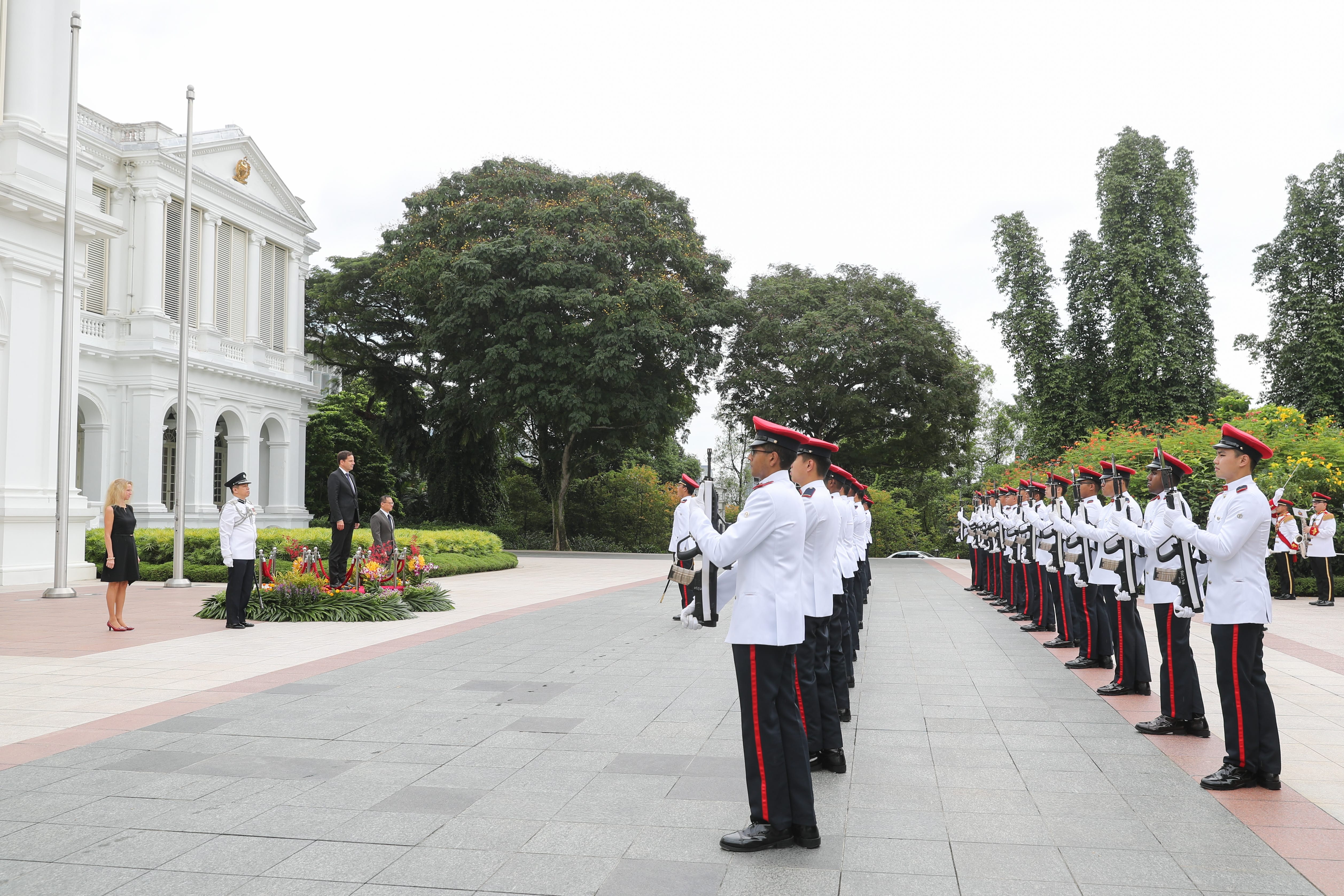Guards in formation, holding rifles, stand at attention near dignitaries in front of a white building.